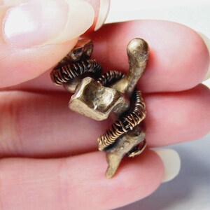 May include: A small, bronze-colored metal sculpture of a snake coiled around a geometric shape. The snake's body is detailed with fine lines, and the sculpture is held between fingers.