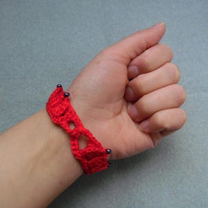 May include: A red crocheted bracelet with black beads. The bracelet is on a person's wrist.