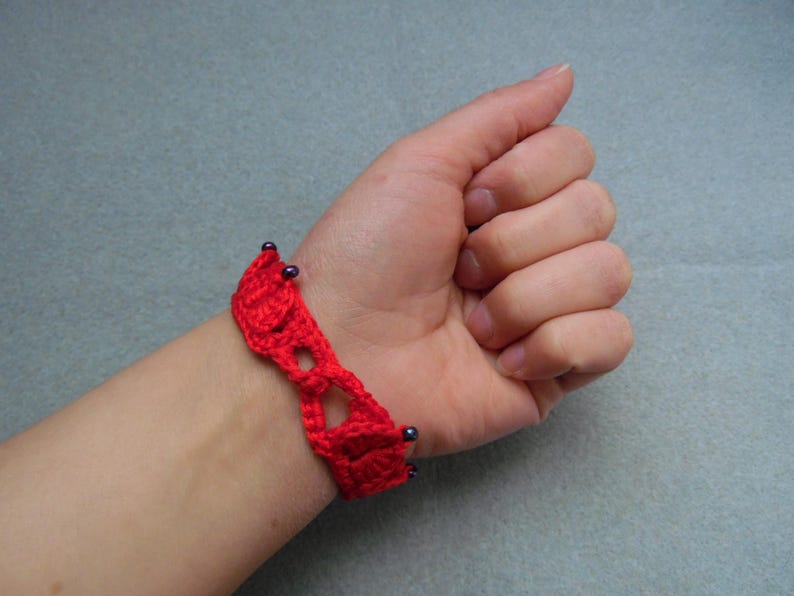 May include: A red crocheted bracelet with black beads. The bracelet is on a person's wrist.