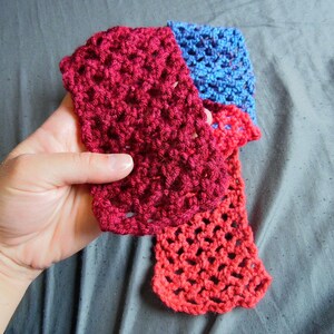May include: A handmade crochet scarf in shades of red and blue. The scarf is held in a hand, showcasing the open, lacy stitch pattern. The colors transition from a deep red to a lighter red and then to blue.