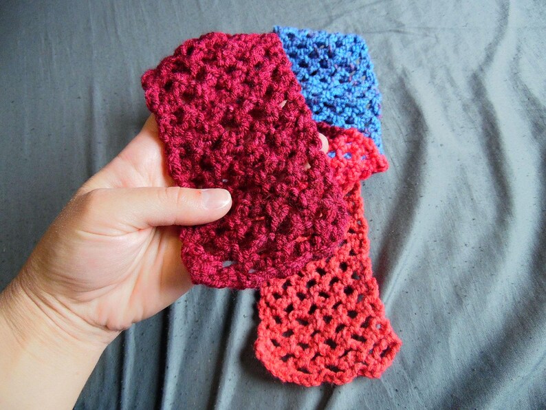 May include: A handmade crochet scarf in shades of red and blue. The scarf is held in a hand, showcasing the open, lacy stitch pattern. The colors transition from a deep red to a lighter red and then to blue.