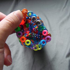 May include: A colorful, handmade beaded bracelet with a cone shape. The bracelet features a variety of vibrant colors, including red, orange, yellow, green, blue, and purple. The beads are connected with silver rings.