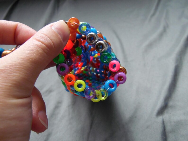 May include: A colorful, handmade beaded bracelet with a cone shape. The bracelet features a variety of vibrant colors, including red, orange, yellow, green, blue, and purple. The beads are connected with silver rings.