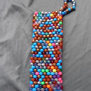 May include: A rectangular beaded item with a loop, crafted from a variety of colorful beads. The beads are primarily blue, brown, silver, and pink, with accents of orange, green, and purple. The item is likely a keychain or accessory.