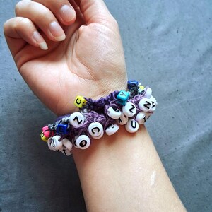 K&ouml;nnte beinhalten: Ein lila geh&auml;keltes Armband, verziert mit wei&szlig;en Buchstabenperlen, die verschiedene Buchstaben buchstabieren. Das Armband hat auch bunte Akzentperlen. Das Armband wird am Handgelenk getragen.