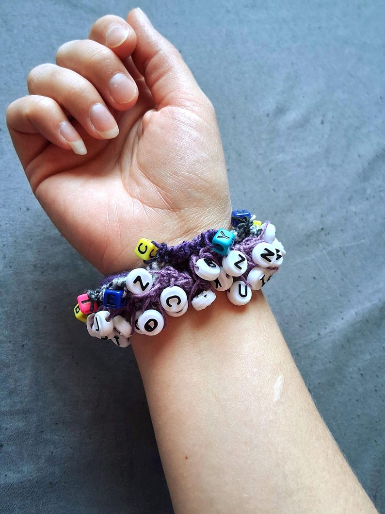 K&ouml;nnte beinhalten: Ein lila geh&auml;keltes Armband, verziert mit wei&szlig;en Buchstabenperlen, die verschiedene Buchstaben buchstabieren. Das Armband hat auch bunte Akzentperlen. Das Armband wird am Handgelenk getragen.