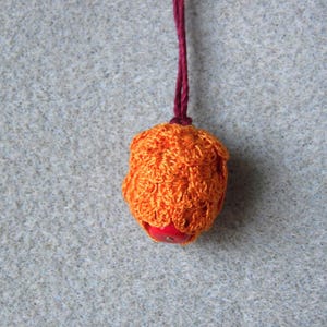 Puede incluir: Adorno de ganchillo naranja hecho a mano con una base de cuentas rojas, suspendido por un hilo burdeos. El ganchillo tiene un diseño texturizado y abierto. El fondo es fieltro gris claro.