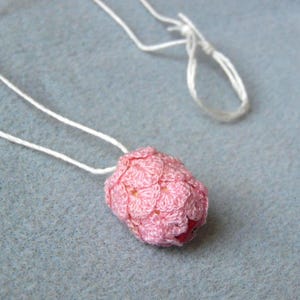 Puede incluir: Un collar con colgante de ganchillo rosa hecho a mano. El colgante tiene forma redondeada con una superficie texturizada, sujeto a un cordón blanco. El collar se muestra sobre una superficie azul claro. El colgante es de color rosa claro.