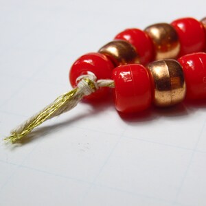 May include: A string of red and copper beads with a gold thread tassel. The beads are strung together with a white thread.