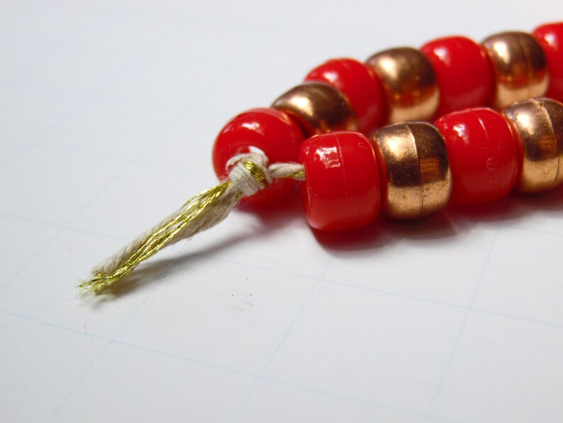 May include: A string of red and copper beads with a gold thread tassel. The beads are strung together with a white thread.