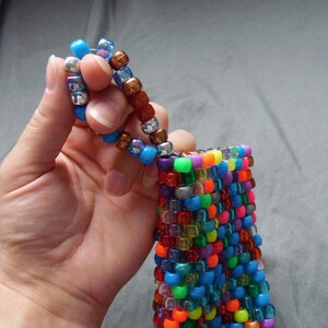 May include: A colorful beaded pouch with a matching beaded handle. The pouch is made of small, square beads in a variety of colors, including blue, green, orange, and purple. The handle is made of similar beads in shades of blue, brown, and silver.