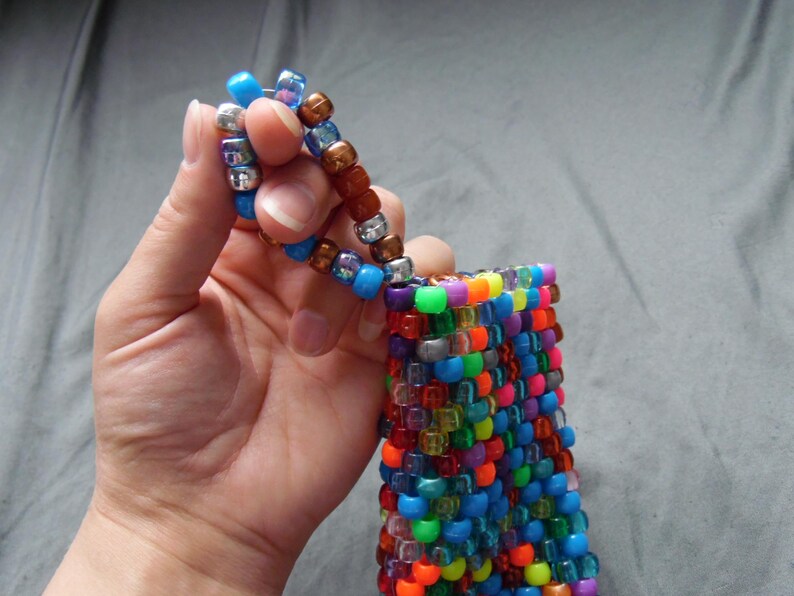 May include: A colorful beaded pouch with a matching beaded handle. The pouch is made of small, square beads in a variety of colors, including blue, green, orange, and purple. The handle is made of similar beads in shades of blue, brown, and silver.
