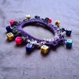 K&ouml;nnte beinhalten: Ein handgefertigtes H&auml;kelarmband mit einem lila Grund und bunten Buchstabenperlen. Das Armband hat eine Mischung aus schwarzem, wei&szlig;em und lila Garn, mit kleinen, quadratischen Perlen in verschiedenen Farben, die Buchstaben buchstabieren. Die Perlen sind um den Umfang des Armbands befestigt.