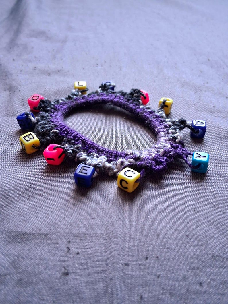 K&ouml;nnte beinhalten: Ein handgefertigtes H&auml;kelarmband mit einem lila Grund und bunten Buchstabenperlen. Das Armband hat eine Mischung aus schwarzem, wei&szlig;em und lila Garn, mit kleinen, quadratischen Perlen in verschiedenen Farben, die Buchstaben buchstabieren. Die Perlen sind um den Umfang des Armbands befestigt.