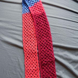 May include: A handmade crochet scarf in shades of blue, coral, and burgundy. The scarf is made with a lacy stitch, creating an open, airy design. The scarf is displayed on a gray surface.