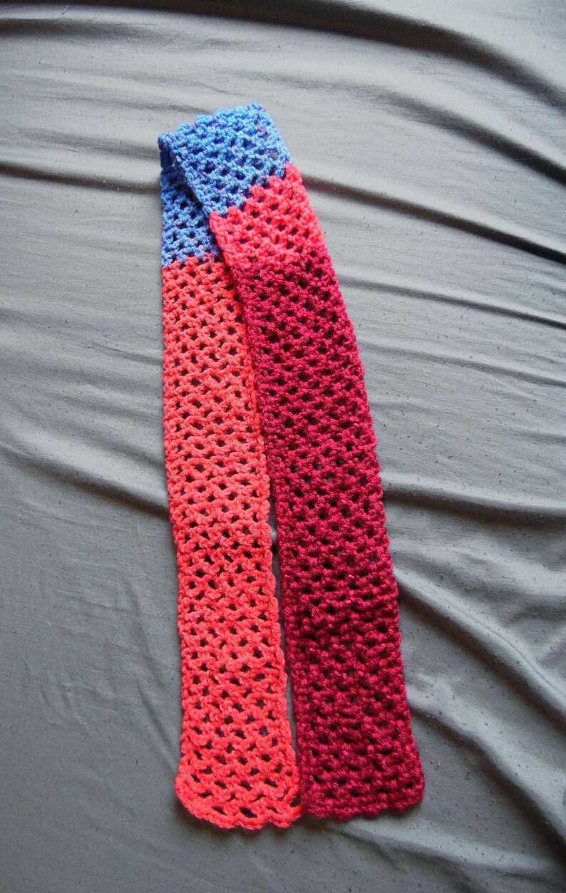 May include: A handmade crochet scarf in shades of blue, coral, and burgundy. The scarf is made with a lacy stitch, creating an open, airy design. The scarf is displayed on a gray surface.