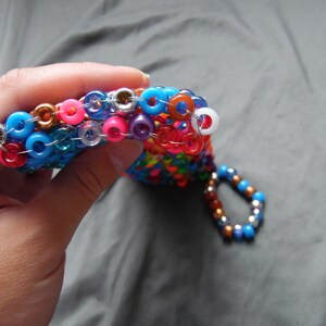 May include: A handmade beaded accessory in a variety of colors, including blue, pink, orange, and purple. The accessory is made of small, round beads strung together with silver-colored metal rings. A small loop of beads hangs from the main piece.