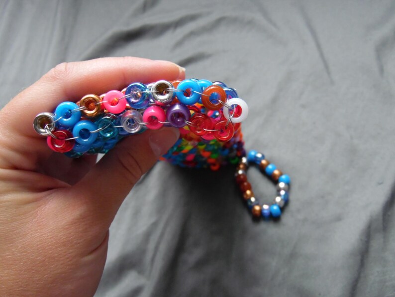 May include: A handmade beaded accessory in a variety of colors, including blue, pink, orange, and purple. The accessory is made of small, round beads strung together with silver-colored metal rings. A small loop of beads hangs from the main piece.