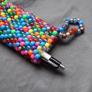 May include: A colorful beaded pen holder with a matching beaded loop. The holder is filled with a black and white pen. The beads are a mix of bright colors including pink, orange, blue, green, and yellow.