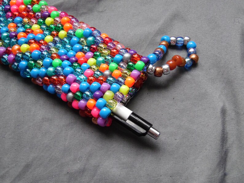 May include: A colorful beaded pen holder with a matching beaded loop. The holder is filled with a black and white pen. The beads are a mix of bright colors including pink, orange, blue, green, and yellow.