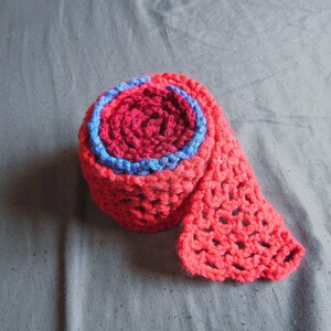 May include: A rolled-up, handmade crochet scarf in shades of red and pink, with a blue accent. The scarf has an open, lacy stitch pattern. The item is displayed on a gray surface.