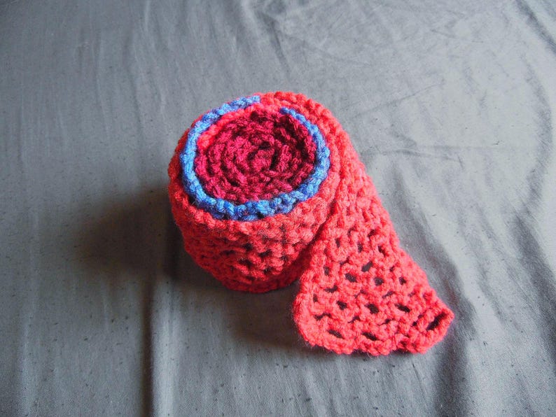 May include: A rolled-up, handmade crochet scarf in shades of red and pink, with a blue accent. The scarf has an open, lacy stitch pattern. The item is displayed on a gray surface.