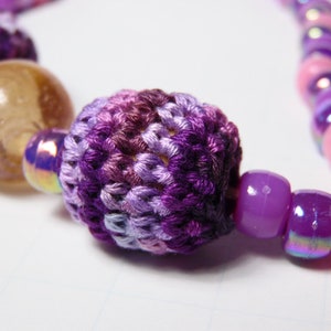 Puede incluir: Un collar con una cuenta de ganchillo de color morado y rosa con cuentas de vidrio iridiscentes de color morado y rosa.