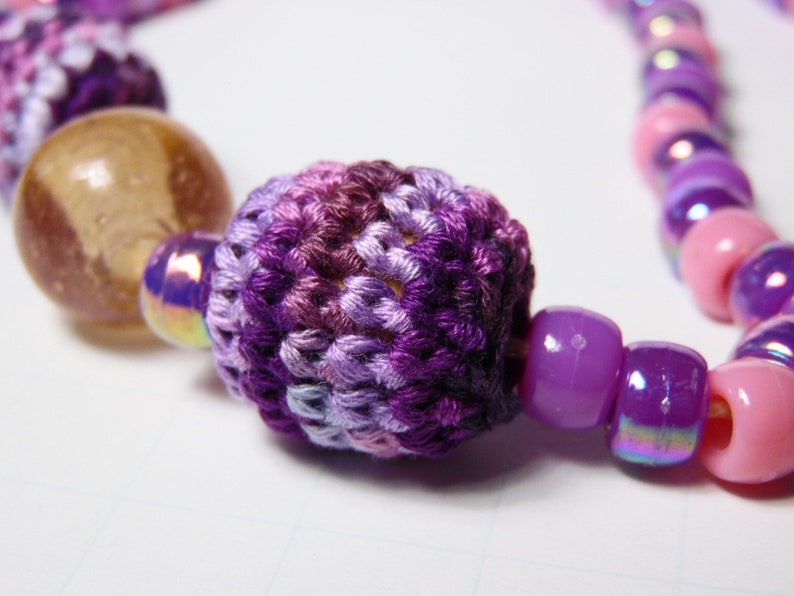 Puede incluir: Un collar con una cuenta de ganchillo de color morado y rosa con cuentas de vidrio iridiscentes de color morado y rosa.