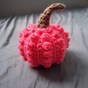 May include: A handmade, bright pink crochet pumpkin with a textured surface. The pumpkin has a brown stem. The craft project is likely for fall or Halloween decor.