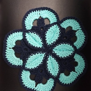 May include: A handmade crochet flower-shaped trivet. The trivet features a navy blue border and light turquoise petals. The trivet is designed to protect surfaces from hot dishes and adds a decorative touch to the kitchen.