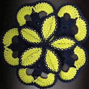 May include: A handmade, crocheted flower-shaped coaster in a vibrant color scheme. The coaster features a dark navy blue base with bright neon yellow-green petals and accents. The design is a circular, floral pattern.