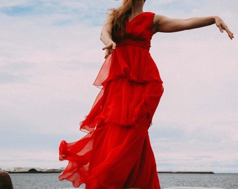 Könnte beinhalten: Ein fließendes, rotes Kleid mit Rüschen. Das Kleid ist ärmellos und hat eine betonte Taille. Das Model steht auf Felsen in der Nähe des Wassers, die Arme ausgestreckt. Der Himmel ist bewölkt mit einem Hauch von Blau.
