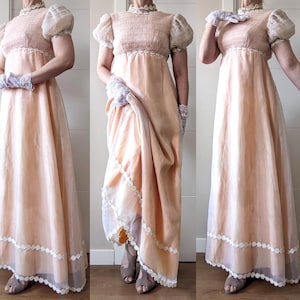 Puede incluir: Un vestido melocotón de cintura imperio con mangas cortas abullonadas y un cuello alto con ribete de encaje. El vestido presenta una falda fluida con un ribete de encaje blanco en el dobladillo. Delicados guantes de encaje completan el conjunto.