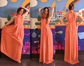 May include: A sleeveless, coral-colored maxi dress with a high neckline and a flowing skirt. The dress features a keyhole back and pleating details. The model is wearing the dress in front of a colorful mural.