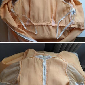 K&ouml;nnte beinhalten: Pfirsichfarbenes Vintage-Kleid mit wei&szlig;em Rei&szlig;verschluss und transparenten &Auml;rmeln. Das Kleid hat eine wei&szlig;e Spitzenborte am Ausschnitt und an den &Auml;rmeln. Das Kleid liegt flach, um seine Details zu zeigen.