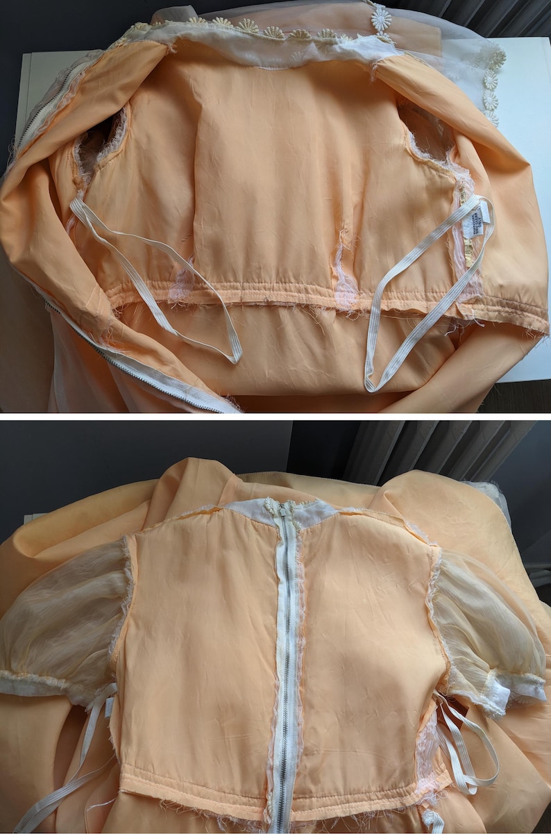 K&ouml;nnte beinhalten: Pfirsichfarbenes Vintage-Kleid mit wei&szlig;em Rei&szlig;verschluss und transparenten &Auml;rmeln. Das Kleid hat eine wei&szlig;e Spitzenborte am Ausschnitt und an den &Auml;rmeln. Das Kleid liegt flach, um seine Details zu zeigen.