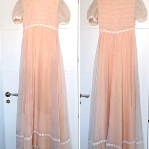 K&ouml;nnte beinhalten: Ein vintage pfirsichfarbenes Kleid mit kurzen, transparenten &Auml;rmeln und hohem Ausschnitt. Das Kleid hat eine zarte wei&szlig;e Spitzenborte am Ausschnitt, an der Taille und am Saum. Das Kleid h&auml;ngt an einem wei&szlig;en Kleiderb&uuml;gel vor wei&szlig;em Hintergrund. Die L&auml;nge betr&auml;gt ca. 150 cm.