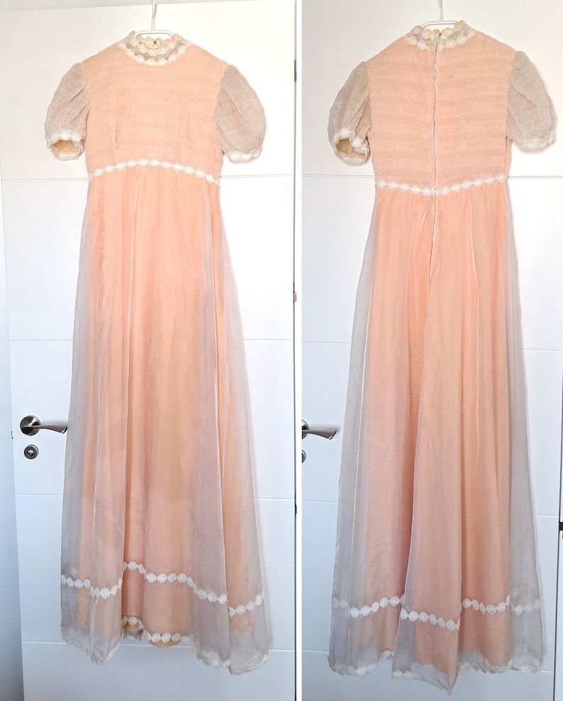 K&ouml;nnte beinhalten: Ein vintage pfirsichfarbenes Kleid mit kurzen, transparenten &Auml;rmeln und hohem Ausschnitt. Das Kleid hat eine zarte wei&szlig;e Spitzenborte am Ausschnitt, an der Taille und am Saum. Das Kleid h&auml;ngt an einem wei&szlig;en Kleiderb&uuml;gel vor wei&szlig;em Hintergrund. Die L&auml;nge betr&auml;gt ca. 150 cm.