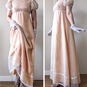 K&ouml;nnte beinhalten: Ein pfirsichfarbenes Empire-Kleid mit kurzen Puff&auml;rmeln und einem hohen, mit Spitze besetzten Ausschnitt. Das Kleid hat einen flie&szlig;enden Rock mit wei&szlig;er Spitzentrimmung am Saum. Das Model tr&auml;gt Spitzhandschuhe und offene Schuhe.