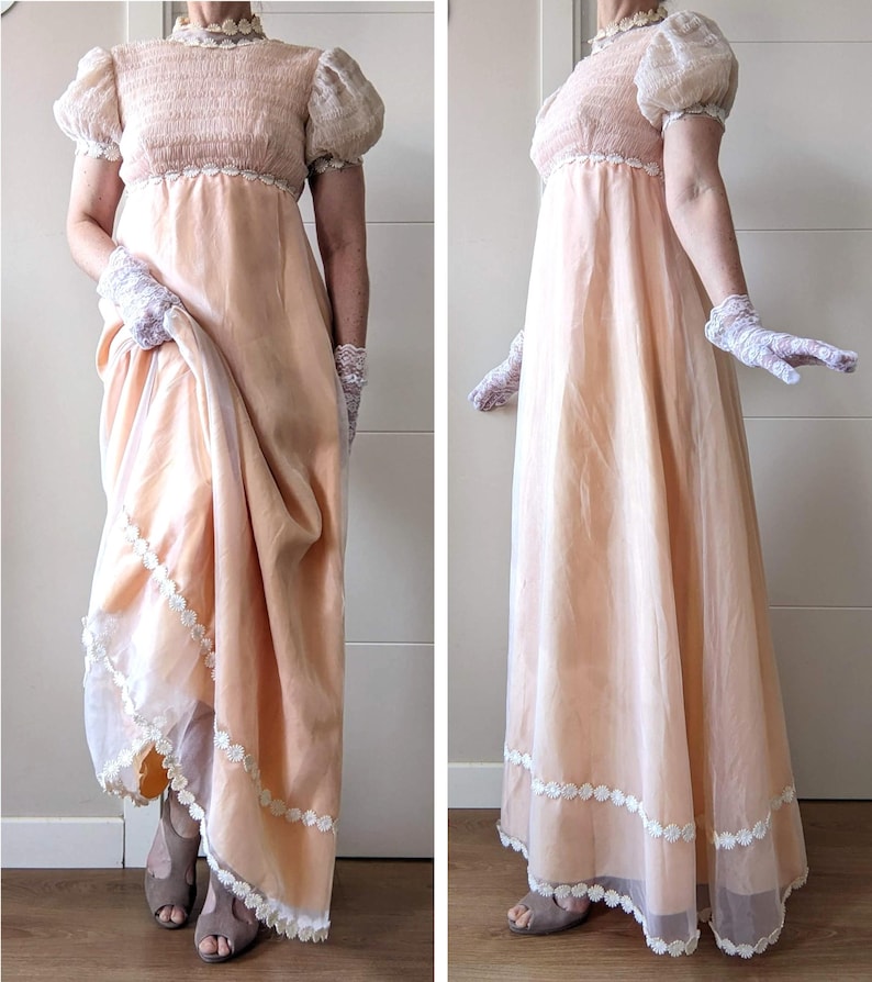 K&ouml;nnte beinhalten: Ein pfirsichfarbenes Empire-Kleid mit kurzen Puff&auml;rmeln und einem hohen, mit Spitze besetzten Ausschnitt. Das Kleid hat einen flie&szlig;enden Rock mit wei&szlig;er Spitzentrimmung am Saum. Das Model tr&auml;gt Spitzhandschuhe und offene Schuhe.
