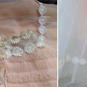 K&ouml;nnte beinhalten: Ein vintage pfirsichfarbenes Kleid mit zarten G&auml;nsebl&uuml;mchen-Details. Das Oberteil hat ein ger&uuml;schtes Design und wei&szlig;e G&auml;nsebl&uuml;mchen-Applikationen. Der Rock hat einen transparenten &Uuml;berzug mit G&auml;nsebl&uuml;mchen-Bord&uuml;re.