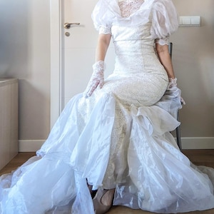 May include: A white floor-length gown featuring intricate lace detailing on the bodice and sleeves, paired with sheer, voluminous organza puff sleeves and a layered organza skirt. The ensemble includes matching white lace gloves with delicate patterns.