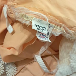 K&ouml;nnte beinhalten: Nahaufnahme eines pfirsichfarbenen Kleidungsst&uuml;cks mit wei&szlig;er Spitzenborte. Ein Etikett lautet "PROFESSIONAL DRY CLEAN ONLY" und enth&auml;lt das ILGWU-Gewerkschaftslabel. Wei&szlig;er Gummizug und Stoff sind sichtbar.