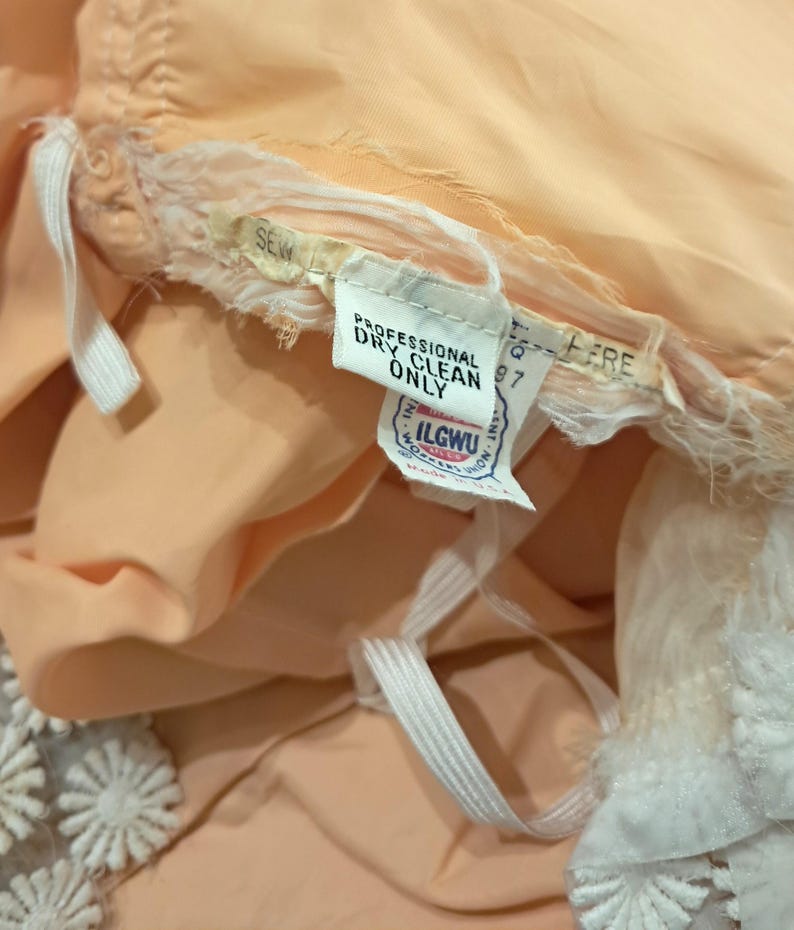 K&ouml;nnte beinhalten: Nahaufnahme eines pfirsichfarbenen Kleidungsst&uuml;cks mit wei&szlig;er Spitzenborte. Ein Etikett lautet "PROFESSIONAL DRY CLEAN ONLY" und enth&auml;lt das ILGWU-Gewerkschaftslabel. Wei&szlig;er Gummizug und Stoff sind sichtbar.