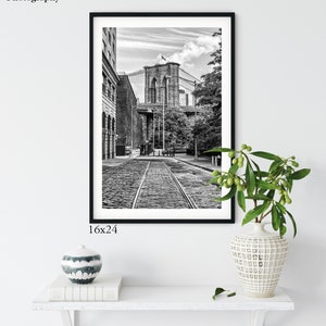 May include: Black and white photograph of the Brooklyn Bridge in New York City. The photo shows a view of the bridge from a street below, with cobblestone streets and a view of the bridge's towers and cables.