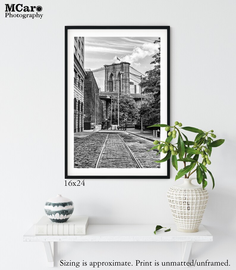 May include: Black and white photograph of the Brooklyn Bridge in New York City. The photo shows a view of the bridge from a street below, with cobblestone streets and a view of the bridge's towers and cables.
