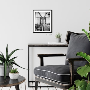 Peut inclure: Photographie en noir et blanc du pont de Brooklyn &agrave; New York. La photo est encadr&eacute;e dans un cadre noir et est expos&eacute;e sur un mur au-dessus d'une table et d'une chaise noires. La taille de l'impression est de 28x36 cm.