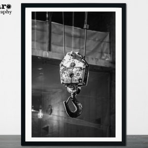 May include: Black and white photograph of a heavy-duty industrial crane hook. The hook is suspended from a cable and is attached to a weathered, metal pulley system. The image is framed in a black border and features the text "MCaro Photography" in the upper left corner.