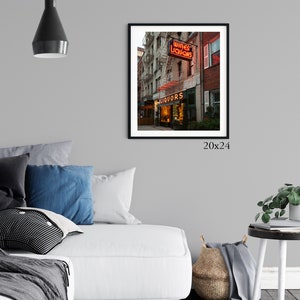 May include: A photograph of a liquor store in New York City with a red neon sign that says "Wines Liquors" and a green awning that says "Liquors". The store is located on a street corner and has a brick facade. The photograph is framed in black and measures 20 inches by 24 inches.