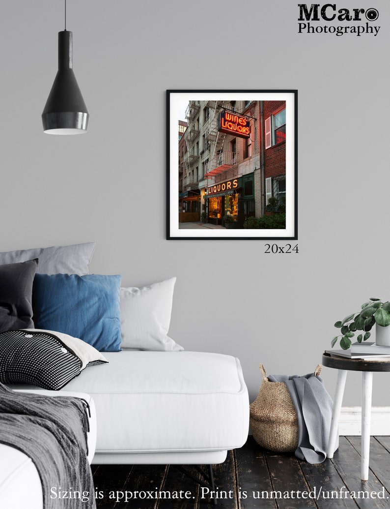 May include: A photograph of a liquor store in New York City with a red neon sign that says "Wines Liquors" and a green awning that says "Liquors". The store is located on a street corner and has a brick facade. The photograph is framed in black and measures 20 inches by 24 inches.